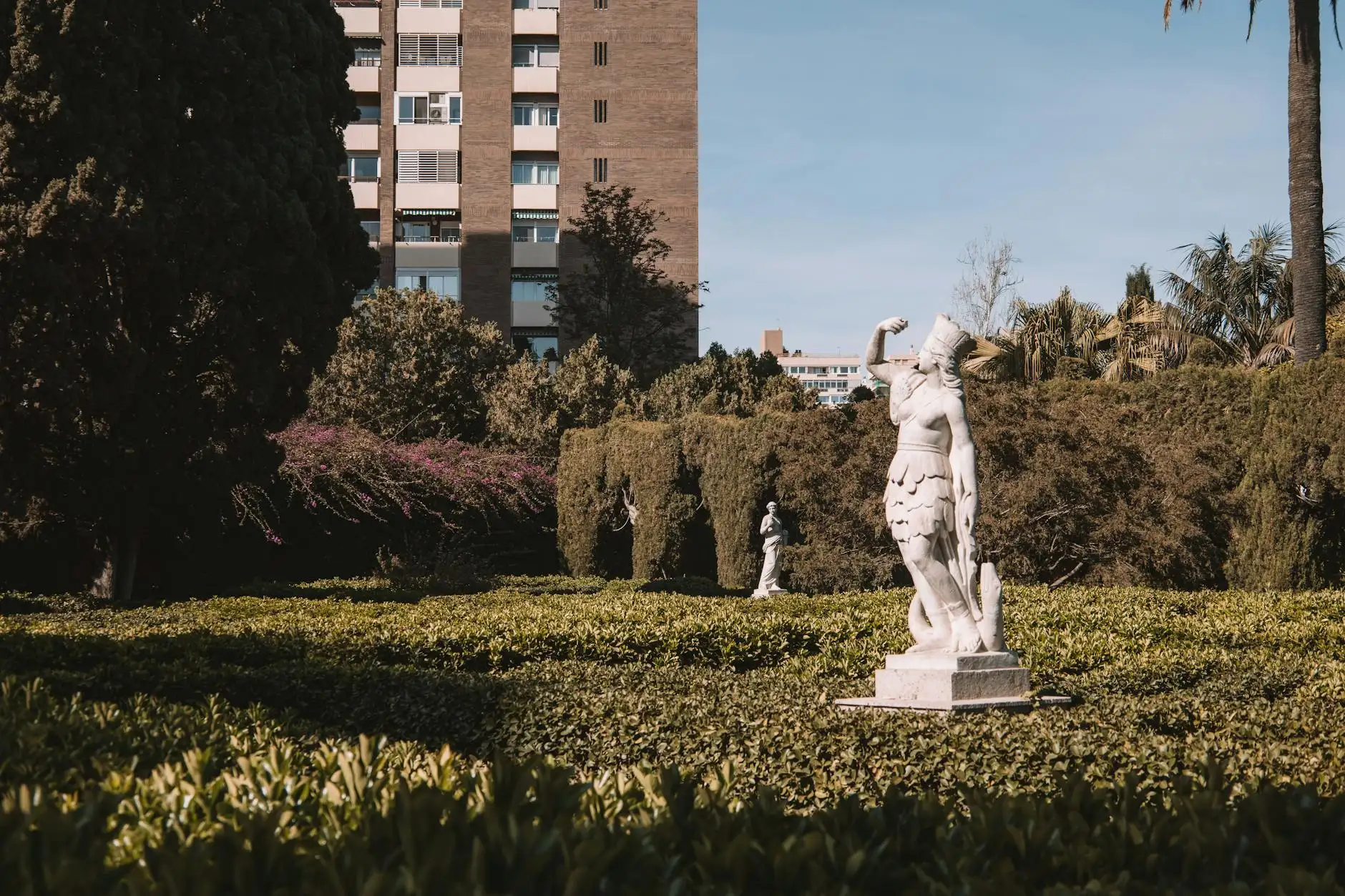 A classical sculpture amidst lush greenery in a Valencia park with urban background.